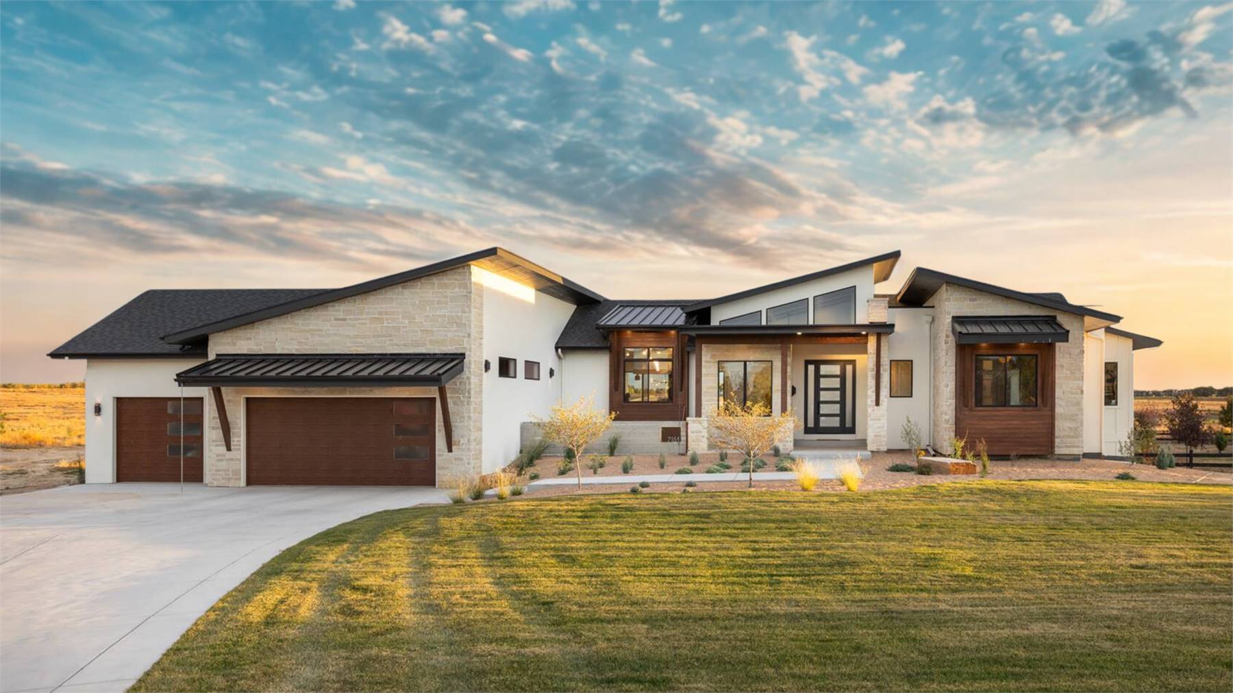 Modern mountain-style home with angled rooflines, mixed stone and wood accents, large picture windows, and a three-car garage at sunset, showcasing House Plan 10833 – Kelowna.